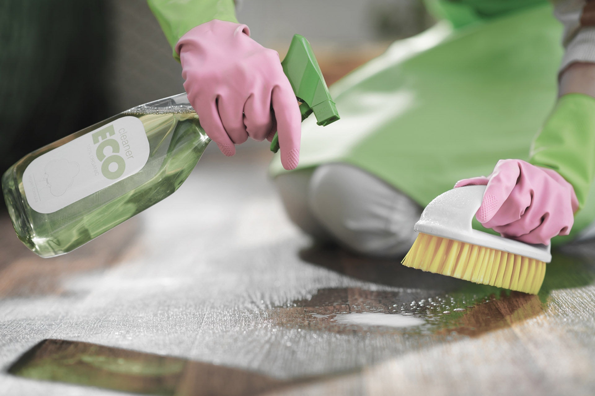 Closeup on woman in green apron and pink rubber gloves with spray bottle of green cleaning supplies and brush spot cleaning floor in the living room in sunny day.